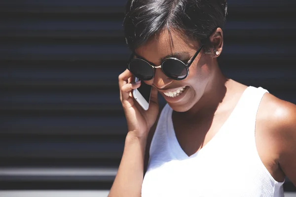 Outdoor portrait of fashionable African American woman in oval sunglasses talking on mobile phone, discussing latest news, smiling, looking cheerful and excited, isolated against dark background