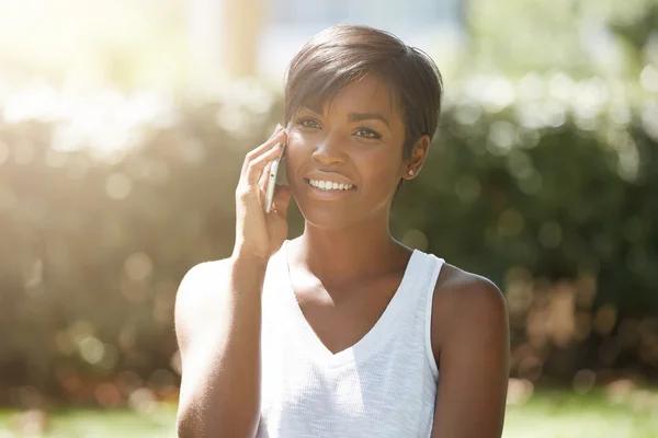 Close up portrait of successful dark-skinned businesswoman dressed casually talking on smart phone to her partners, smiling, joking, having a nice talk outdoors against public garden background