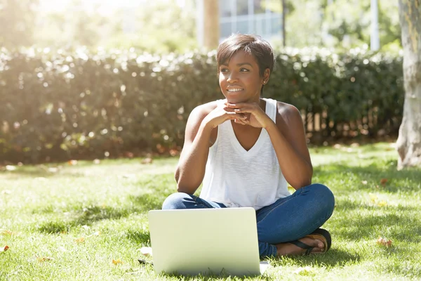 Human and technology concept. Charming African woman with short pixie hairstyle enjoying sunny weather, sitting on the lawn in front of laptop computer in the public garden, waiting for her friend