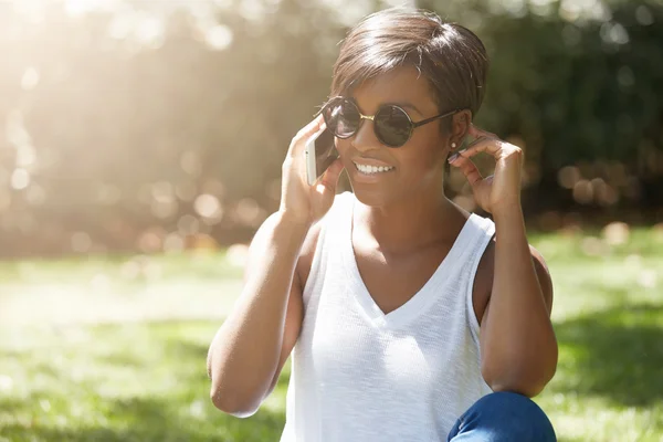 Headshot of stylish young woman with short pixie haircut wearing round shades making phone calls, smiling and flirting while having a conversation with her boyfriend, sitting on the green grass