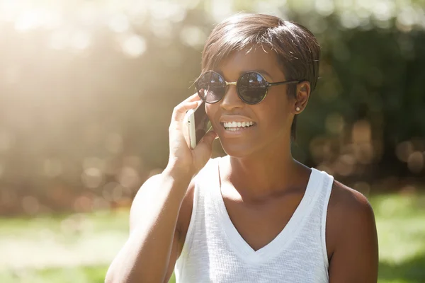 People, technology and communication concept. Headshot of cute African American young woman with short hairstyle wearing hipster shades, smiling while speaking on cell phone in the public garden