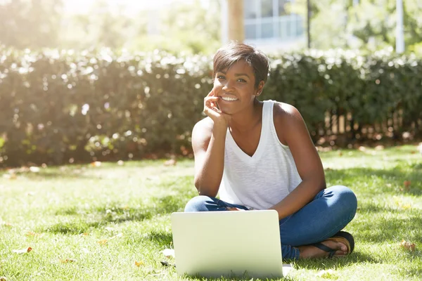 Happy young black female traveller, sitting on the grass, chatting with her family using laptop, smiling widely looking at the camera against public garden background. People and technology concept