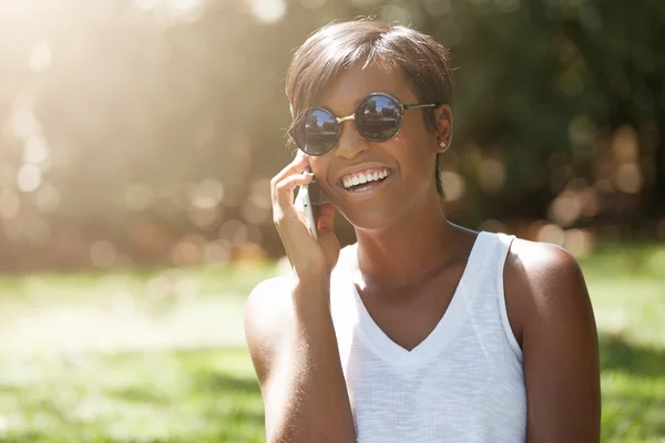 Close up portrait of dark skinned female laughing at a joke while having a phone conversation, enjoying summer sunny day in the city park, wearing stylish sunglasses, looking carefree and happy