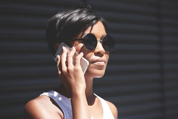 Worried black female with short haircut wearing round shades calling her boyfriend on cell phone trying to find him in a crowded street standing against blank wall with copy space for your information