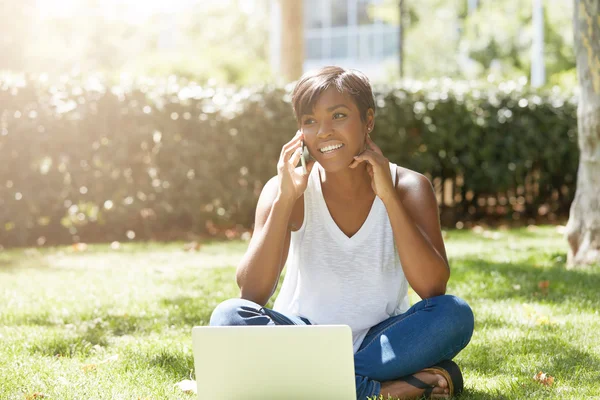Outdoor portrait of black stylish female sitting on the grass in the park, talking on the phone, order a table for evening. Smiling African girl using laptop computer outdoors, touching her ear