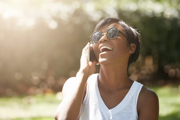 Human face expressions and emotions. Fashionable young dark-skinned female wearing stylish sunglasses having a nice phone talk, smiling, laughing with mouth wide open relaxing in the urban park
