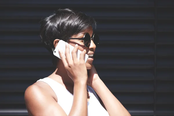 Portrait of smiling good-looking African female in round hipster sunglasses looking puzzled and confused describing her location to a friend on cell phone. Technology and communication concept