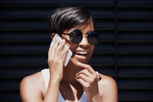 Outdoor portrait of fashionable African American woman in round shades talking on smart phone to her friend, discussing latest news, smiling, laughing, looking cheerful and excited, touching her chin