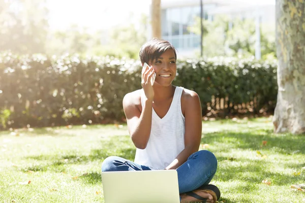 Young charismatic black female business coach sitting on the grass in the park using laptop, talking on mobile phone with asmile, waiting for her trainees to come for training activity outdoors