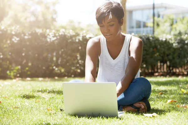 Young attractive dark-skinned college student wearing tank top and jeans sitting on the lawn at campus on sunny day, working on her thesis using laptop computer, looking busy and concentrated