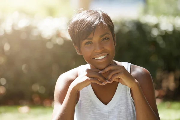 Cropped portrait of stylish charismatic dark-skinned female dressed casually posing with happy smile and hands on her chin, sitting on the green lawn against blurred background of green nature