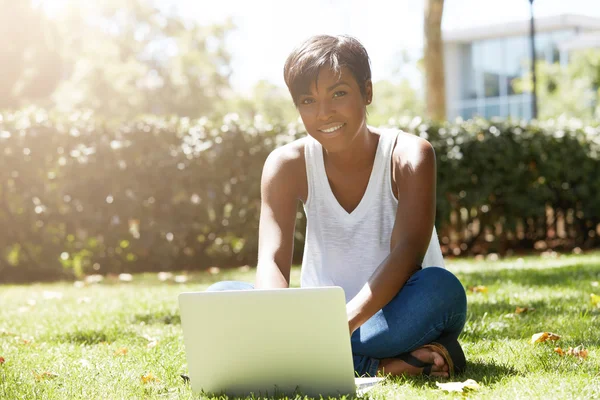 Young pretty African female freelancer dressed casual, sitting on in the sun on the lawn in front of notebook, working on her laptop computer, translating articles, looking at the camera with a smile