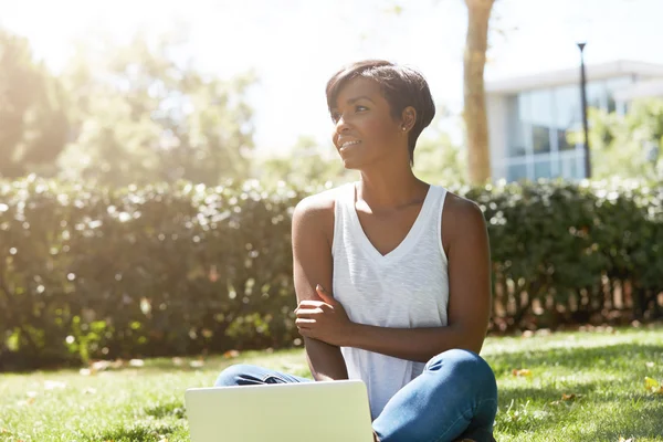 Outdoor portrait of young charming African American woman, sitting on the grass at a park with her laptop, dreaming, looking aside with a smile, having closed posture with thoughtful face expression