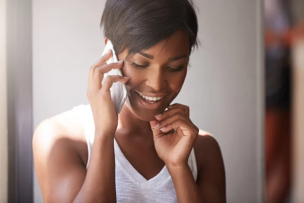 Close up portrait of beautiful dark skinned woman with healthy skin talking on cell phone with her man, smiling, looking happy, shy, and embarassed, touching her cheek, against white wall background