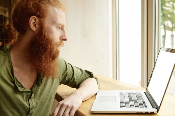 Indoor profile of young extraordinary redhead bearded Caucasian man with serious face expression working on his laptop computer with blank copy space screen for your text or advertising content,