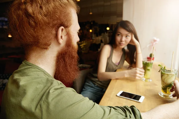 Rear view of stylish Caucasian man with beard sitting at cafe table with drinks and copy space mobile phone on it, having nice dialogue with his good-looking brunette Asian girlfriend during date