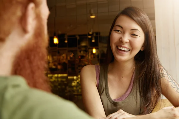 Young beautiful Asian student girl with happy and cheerful expression on her face talking to her redhead groupmate with beard, laughing at his jokes during meeting at cafe after lectures at university