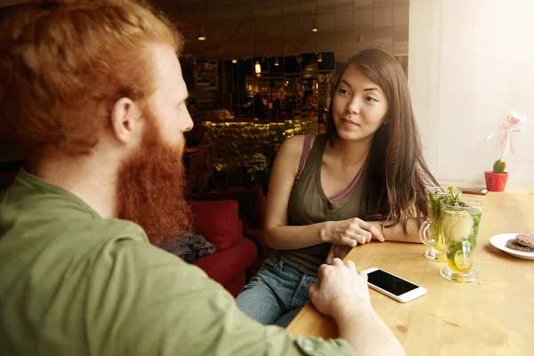 Young entrepreneurs discussing common project sitting at coffee shop, ordered mint tea. Asian woman having concerned look, Caucasian man touching his mobile with blank space for your advertisement