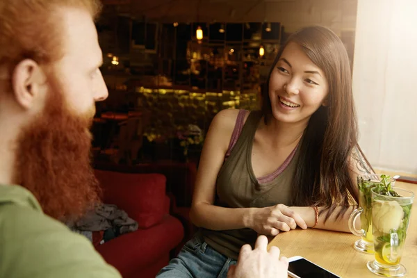 Young attractive Asian girl with dark hair having conversation with redhead stylish-looking man, laughing, having fun, drinking fruit cocktails. Cute multhiethnic couple on date at cozy restaurant
