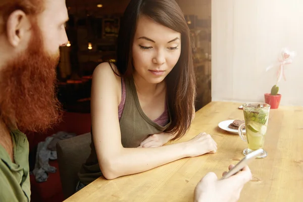 Interracial couple having fresh drinks at coffee shop. Caucasian bearded man talking on his start up project, holding cell phone, showing pictures to his Asian friend who is looking at screen