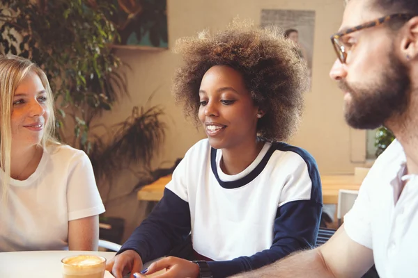 Old friends meeting at cafeteria: young happy dark-skinned girl with Afro haircut having nice conversation with blonde female and bearded man in glasses, telling some funny story of her life