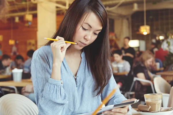 Attractive Asian student girl wearing casual blouse sitting at university canteen with electronic tablet, browsing Internet and making home task, holding pencil at her cheek with pensive expression
