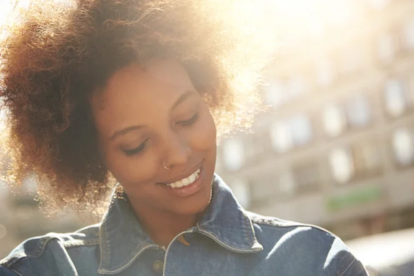 Attractive happy dark-skinned model with Afro hairstyle and nose-ring, posing outdoors against urban background during her morning walk, looking down with shy smile showing her white teeth. Flare sun