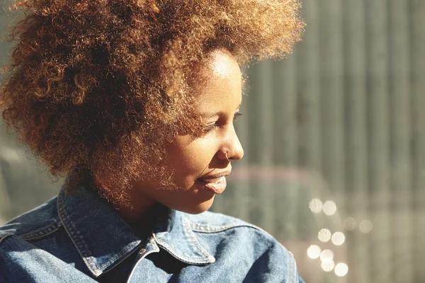 Outdoor portrait of young fashionable African American female with stylish curly hair and ring in her nose, wearing denim jacket, looking away, standing at wall with copy space for your information