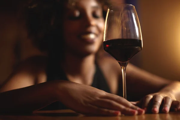 Cropped shot of dark skinned female touching glass of red wine with both hands, selective focus on drink. Black woman with Afro hairstyle sitting at bar, smiling, waiting for friend to join her later
