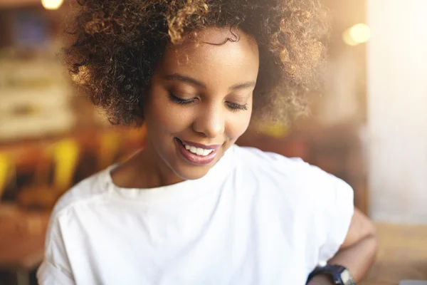 Close up of good-looking African woman with curly hair dressed in white t-shirt, spending free time at cafeteria, looking down with shy cute smile, showing her white teeth, against blurred background