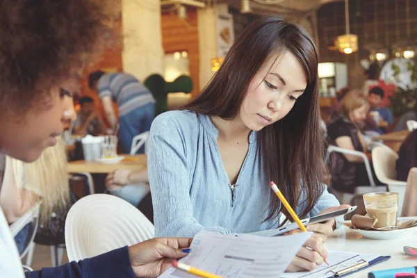 Two female students doing home task at cafe, having coffee. Asian girl holding touch pad, writing out data in notebook while her colleague studying graphics, making notes and filling in some papers