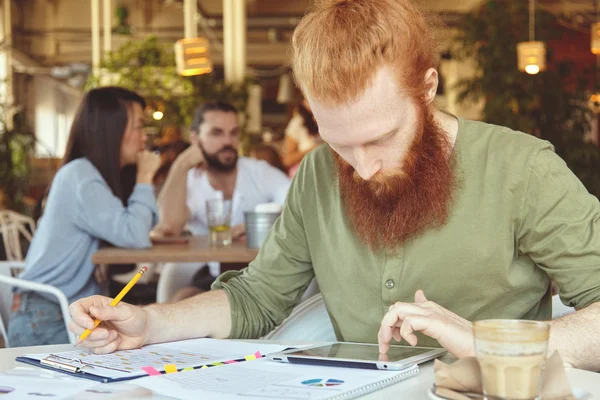 Young trendy entrepreneur with long red hair and beard working on prospective business plan sitting at coffee shop, reading something at pad, filling in charts on sheet of paper with another hand