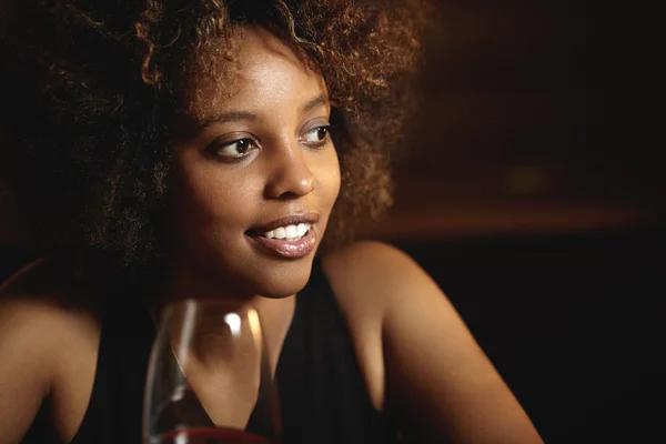 People and leisure. Elegant African woman with perfect tanned skin sitting at restaurant, holding glass of red wine, looking pensive and mysterious. Attractive female spending nice time at bar