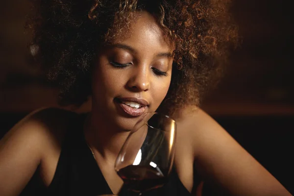 Portrait of young attractive dark-skinned charming woman with curly hair wearing black elegant dress, enjoying delicious red wine, celebrating New Year\'s eve with her friends at bar or restaurant