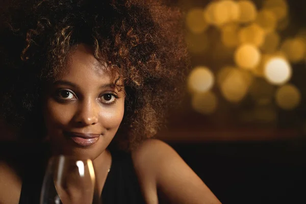 Close-up portrait of smiling black woman with healthy skin relaxing at bar counter, holding glass of wine, looking at camera with happy brown eyes, against blurred background of New Year decoration