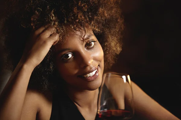 Portrait of gorgeous dark-skinned woman with Afro hairstyle touching her forehead at New Year celebration party at nightclub with glass of wine, smiling at camera with shy mysterious face expression