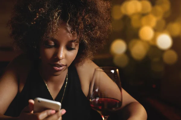 Chic dark-skinned female with Afro haircut, chilling at bar, drinking red wine, sitting at counter, holding mobile phone, texting friends via social networks. African girl typing message at night club