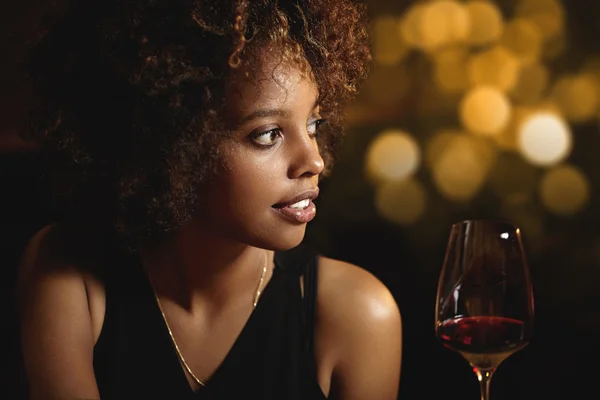 Profile of beautiful African woman with stylish curlly hair wearing elegant black dress, enjoying red wine, siting at restaurant, having romantic dinner with husband, celebrating wedding anniversary