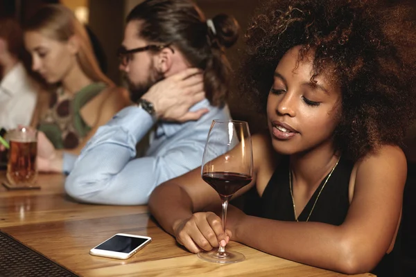 Sad pensive down hearted dark-skinned woman with glass of red wine at bar counter during New Year celebration, looking at cellphone with copy space for your advertisement, waiting for important call