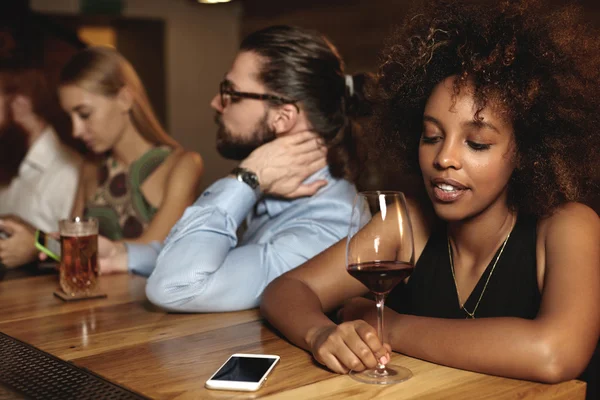 Young people sitting and chatting at bar at New Year party, drinking beverages. Selective focus on couple of diverse races, black girl looking at glass of wine, Caucasian man surfing mobile Internet