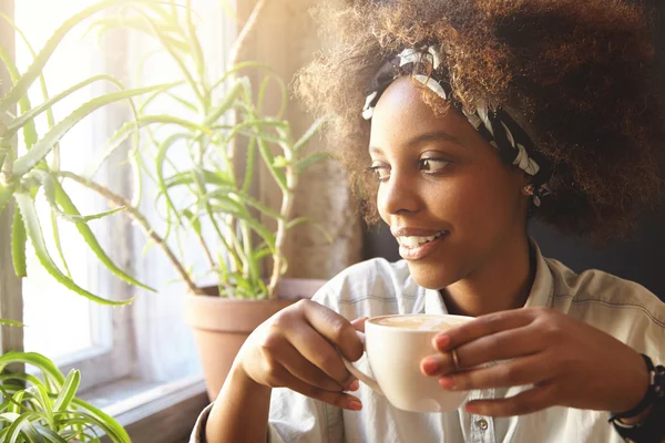 People and leisure. African woman dressed in trendy clothes holding cup of coffee or tea, enjoying hot drink. Black student girl having rest at cafe after college, drinking cappuccino, looking relaxed