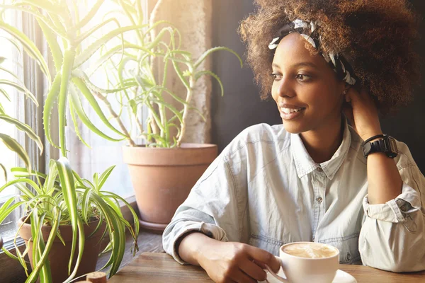 Fashionable African girl with do-rag on her head sitting at cafe table, enjoying morning cappuccino. Young black woman drinking coffee, contemplating urban views through window with happy smile