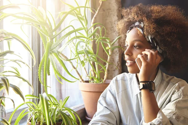 Cute African female with stylish curly haircut having nice conversation with her friend using gadget, making appointment, listening attentively. Young woman sitting at cafe, talking on cell phone