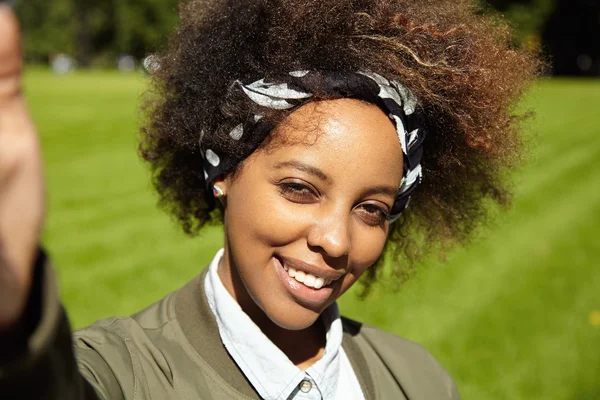 Young stylish woman with Afro haircut wearing black bandana, taking selfie, holding mobile phone or other device in right hand, smiling and squinting eyes in bright sun, standing on green lawn