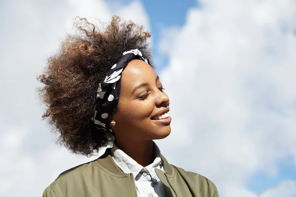 Profile of young African girl having walk in open air, smiling with closed eyes, wind playing in her curly hair. Pretty woman with happy smile, dreaming and enjoying good sunny weather outdoors