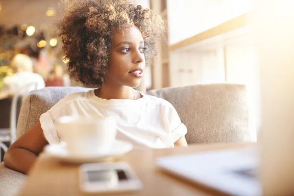Selective focus. Headshot of serious black woman with Afro hairstyle, dressed in casual white top, sitting on sofa at cafe and waiting for her friend. African student using laptop and having coffee