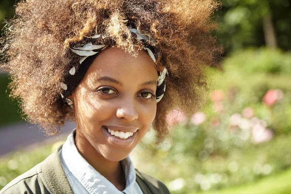 Happy stylish African girl with curly hair enjoying sunny weather in urban garden, breathing fresh air, looking at camera with joyful and pleased smile. Young woman resting outdoors on sunny day