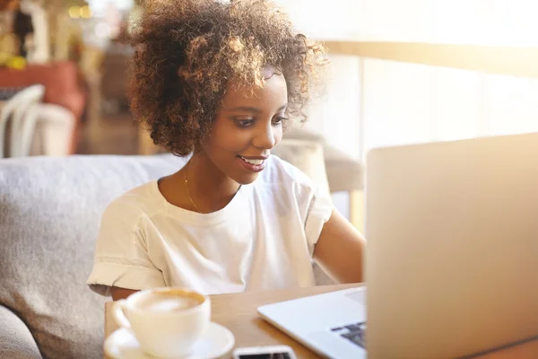 Good-looking dark-skinned female fashion blogger with stylish curly hair, typing post on new trends using laptop. looking at screen with inspired smile, sitting at cafe table with mug and mobile phone