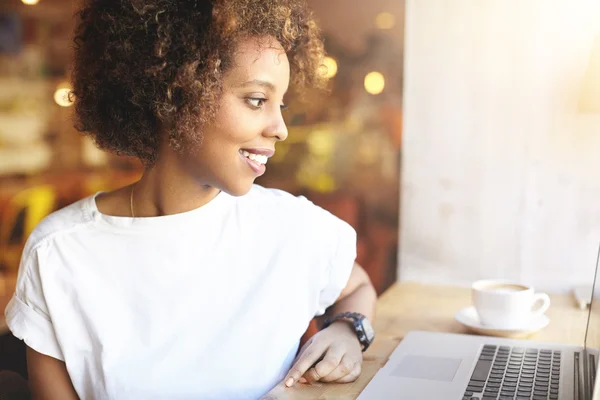 Student girl browsing Internet, using free wi-fi at cafe. African freelancer thinking on ideas for her blog, using laptop at co-working space, resting hand on wooden table, looking with inspired smile