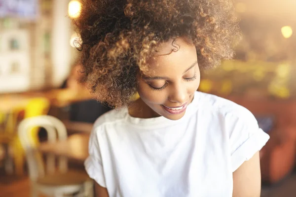 Beautiful dark-skinned student girl with curly hair, dressed in white casual top, waiting for her college mate at cafeteria for lunch, looking down with cute shy smile against blurred background